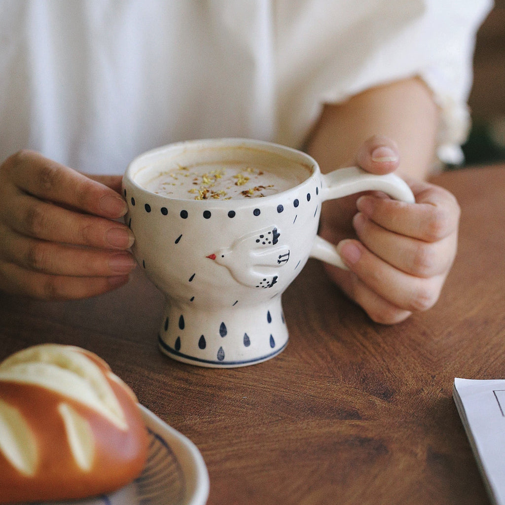 Person holding a polka dot mug with a cat design on a wooden table.