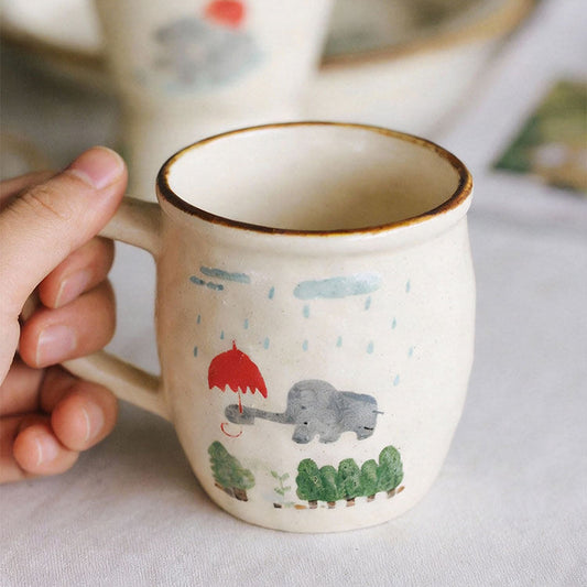 Ceramic mug with animal illustrations held by a hand on a light surface.