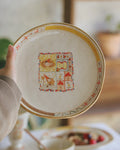 Decorative plate with a colorful design held by a hand, blurred background of a table setting.