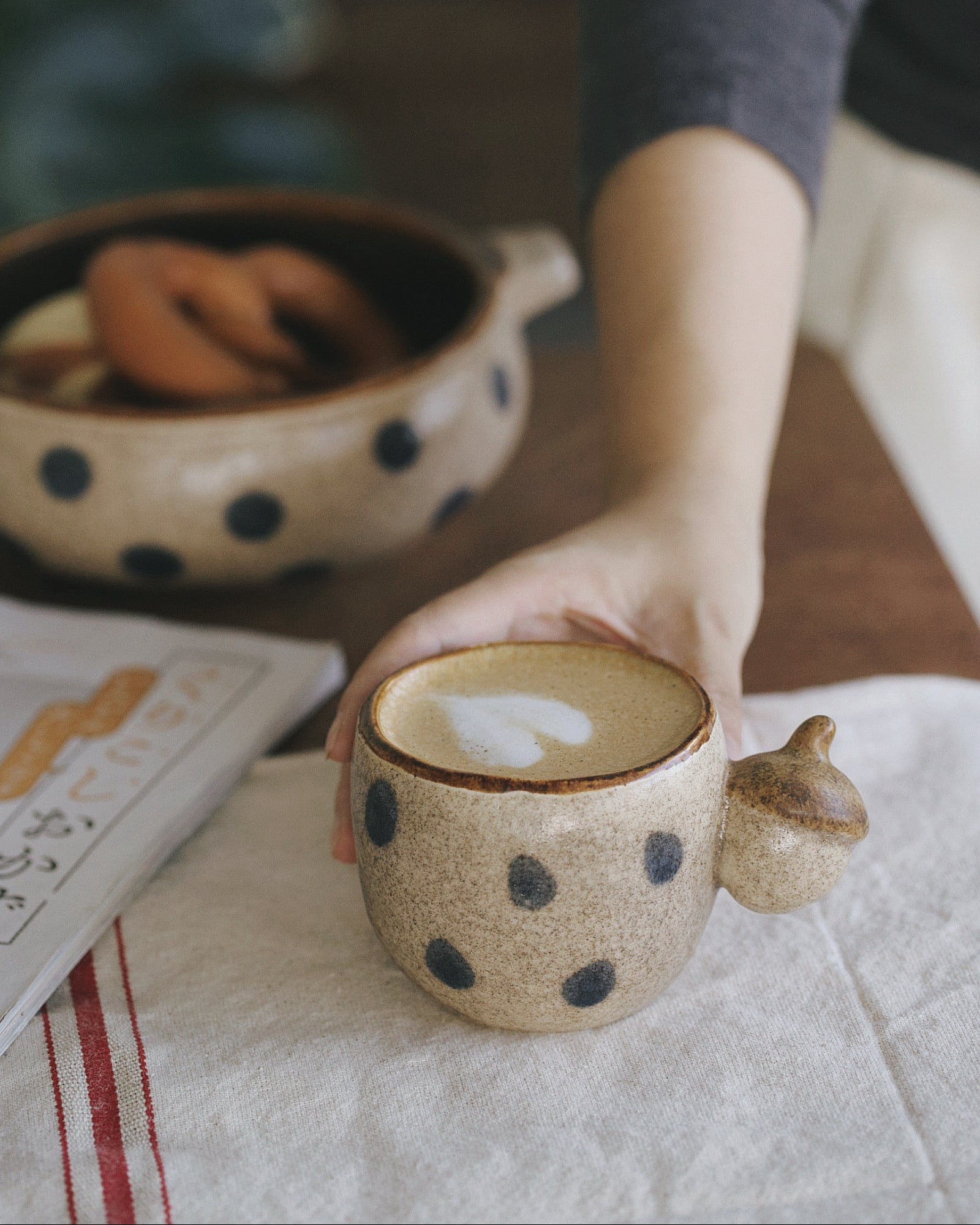 Person holding a ceramic mug with polka dot design on a table with a book and bowl in the background.
