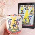 Mug with artistic design of people on a stage held by a hand, with a tablet displaying the same design in the background.