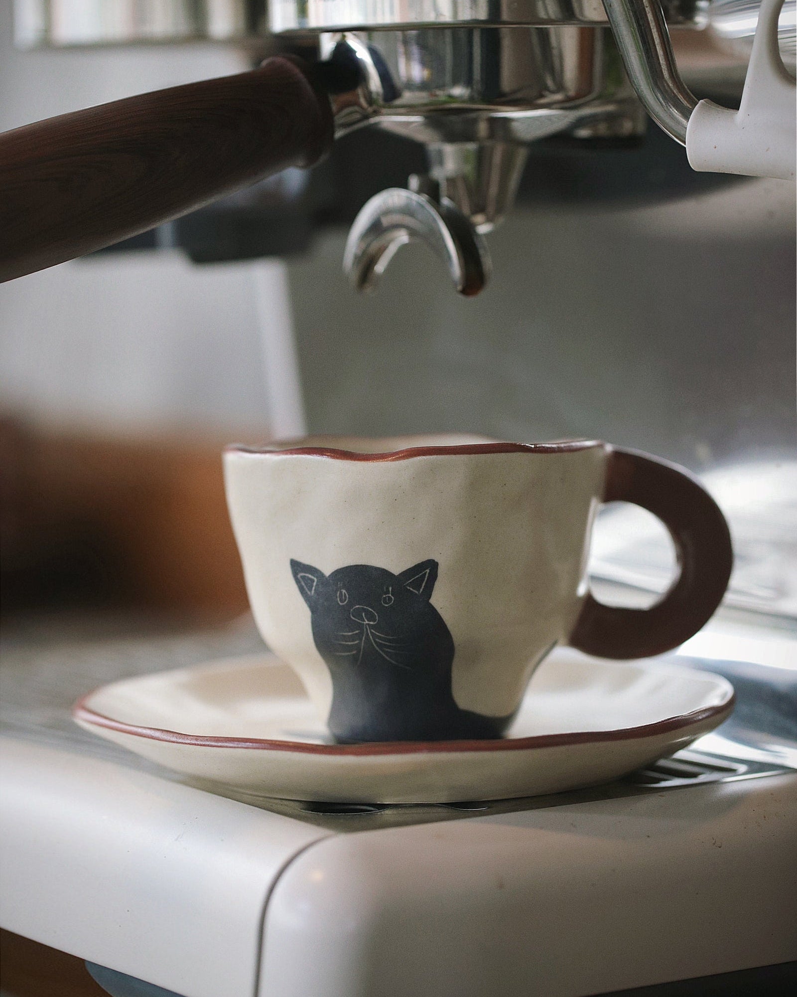Espresso being poured into a cup with a cat design on a saucer, next to an espresso machine.