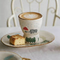 Ceramic cup with a design of a red umbrella and gray hippo, placed on a saucer with a piece of bread.