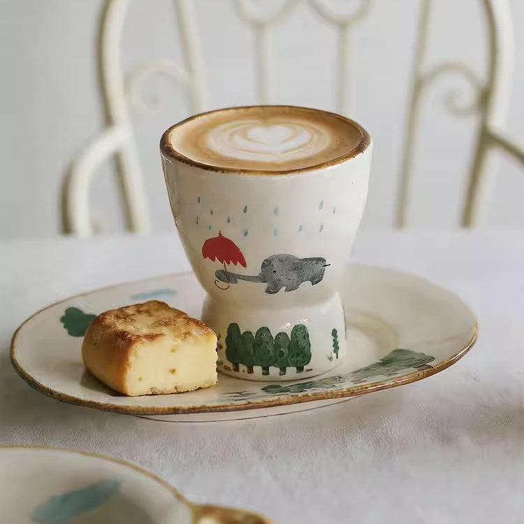 Ceramic cup with a design of a red umbrella and gray hippo, placed on a saucer with a piece of bread.
