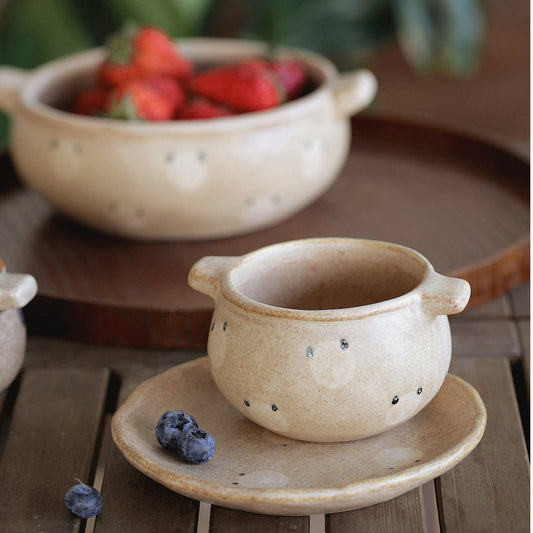 Ceramic cup and saucer on a wooden surface with a bowl of strawberries in the background.