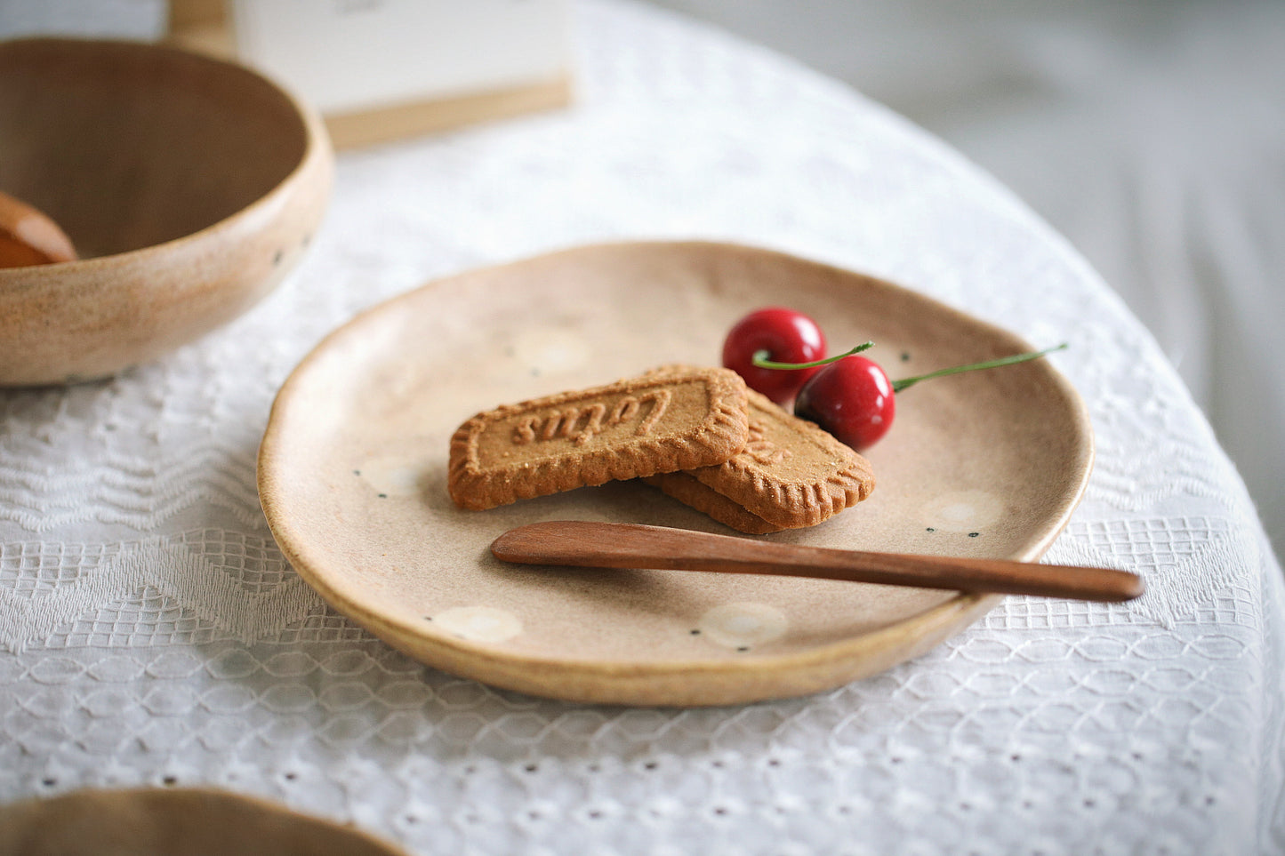 Two cookies on a ceramic plate with a wooden spoon and two red berries on a white lace tablecloth.