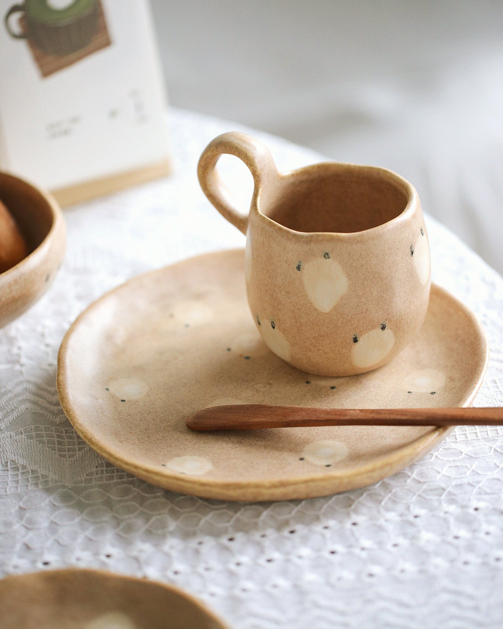 Ceramic mug with a face on a plate with a wooden spoon on a white tablecloth.