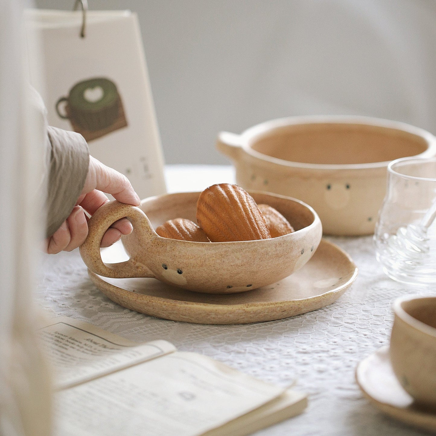 Person holding a ceramic bowl with bread on a table with other ceramic items.