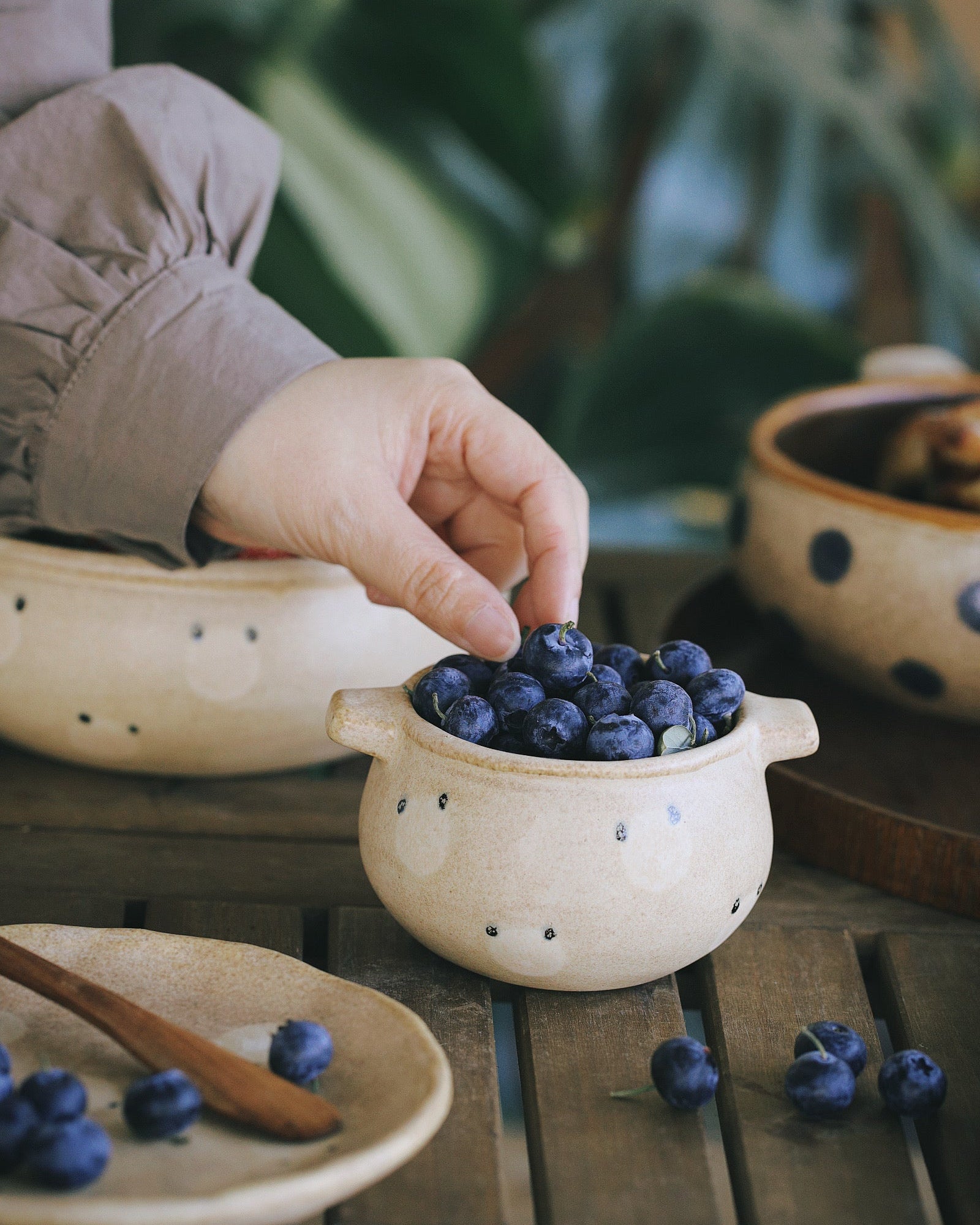 Hand picking blueberries from a small ceramic bowl on a wooden table with a blurred green plant background.