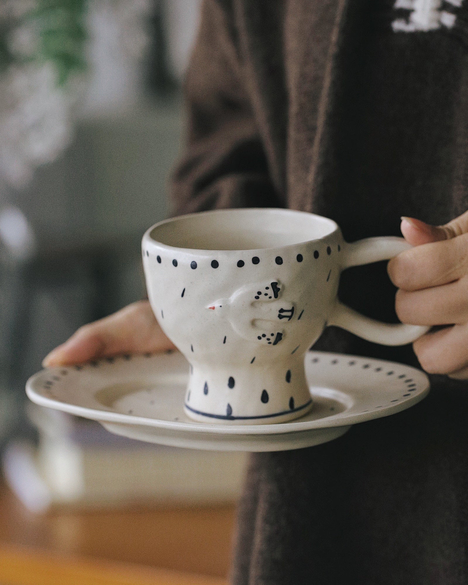 Person holding a polka dot mug and saucer in a blurred indoor setting
