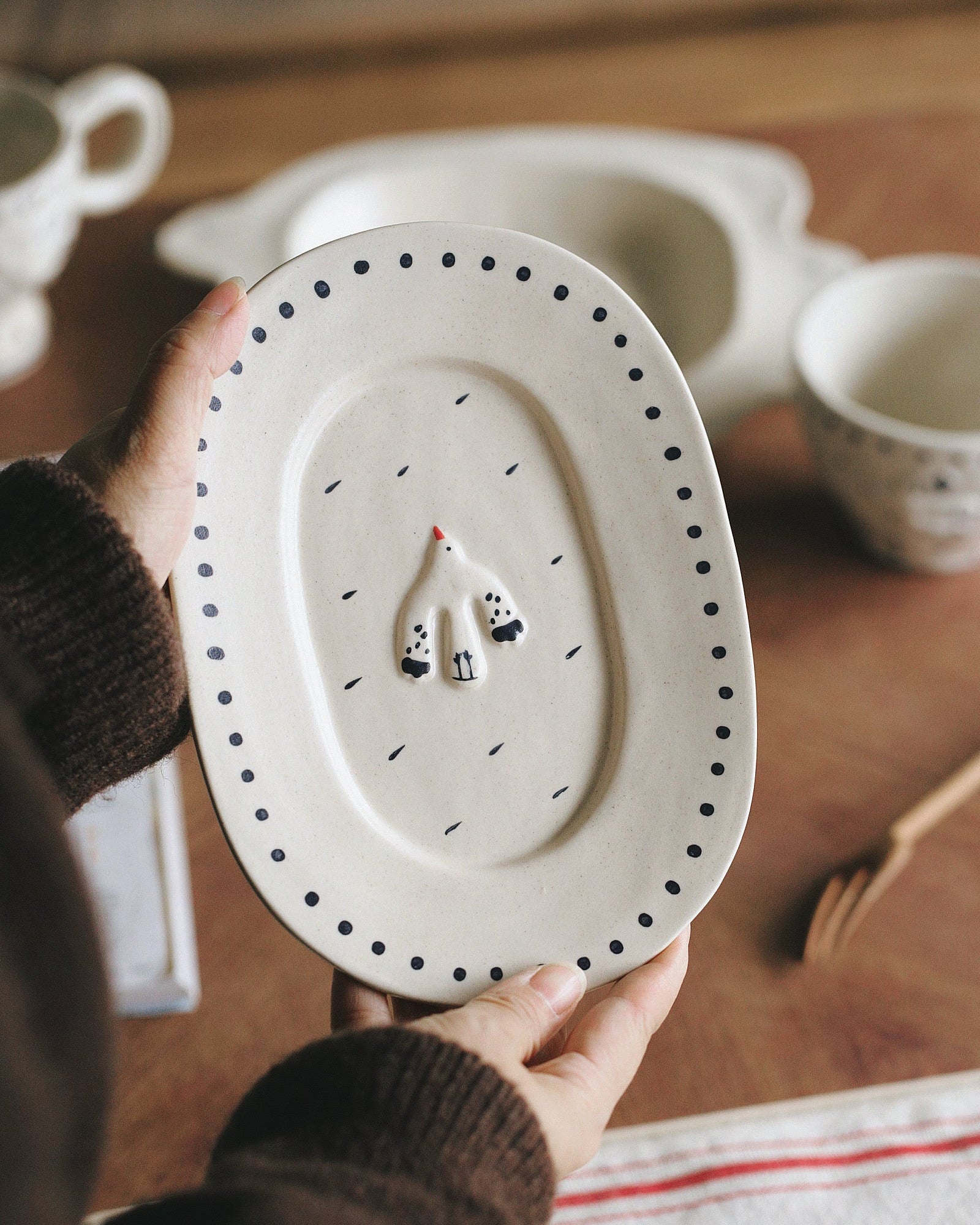 Person holding a ceramic plate with black dots and small figures on a wooden surface.