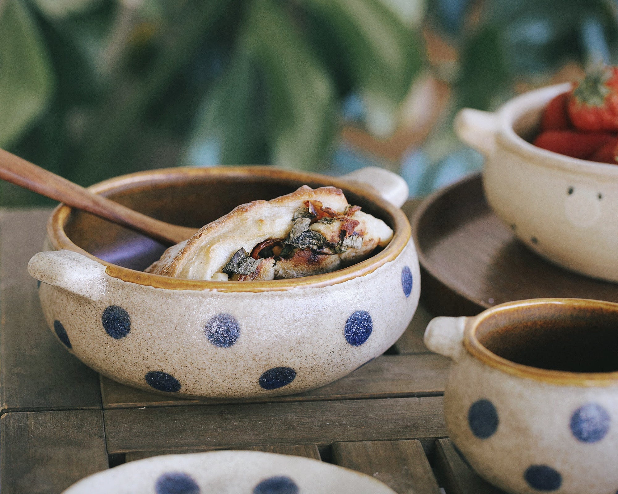 Ceramic bowl with blue polka dots containing food, wooden spoon, on a wooden surface with blurred greenery in the background.