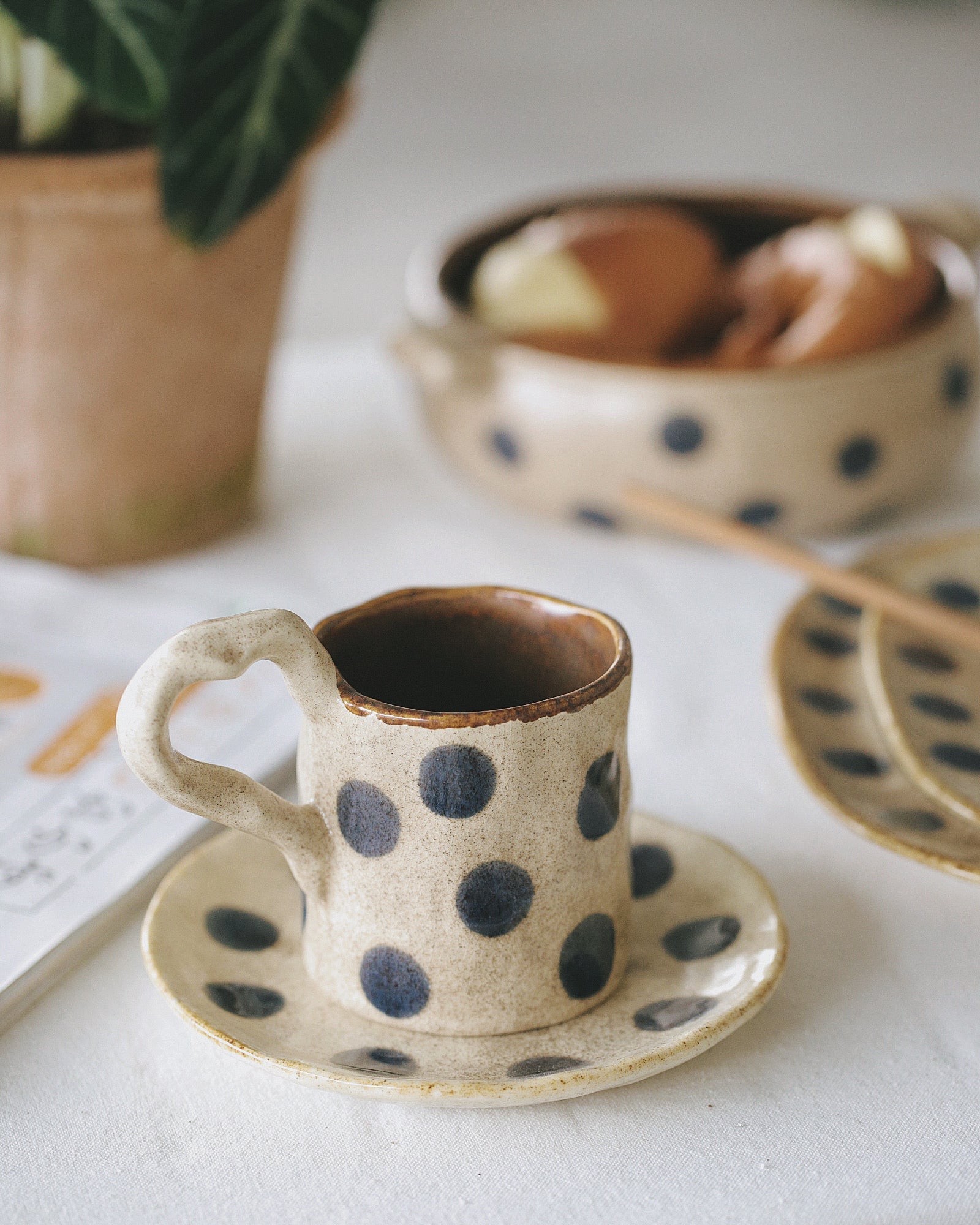 Ceramic cup and saucer with blue polka dots on a light surface, with a bowl and plant in the background.