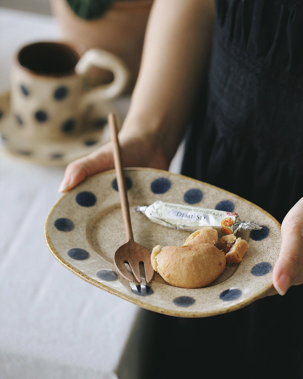 Person holding a plate with a pastry, fork, and tube of cream next to a cup of coffee.