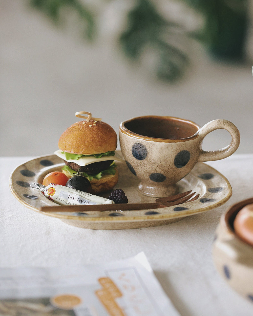 Small burger and polka dot cup on a plate with a blurred green plant in the background