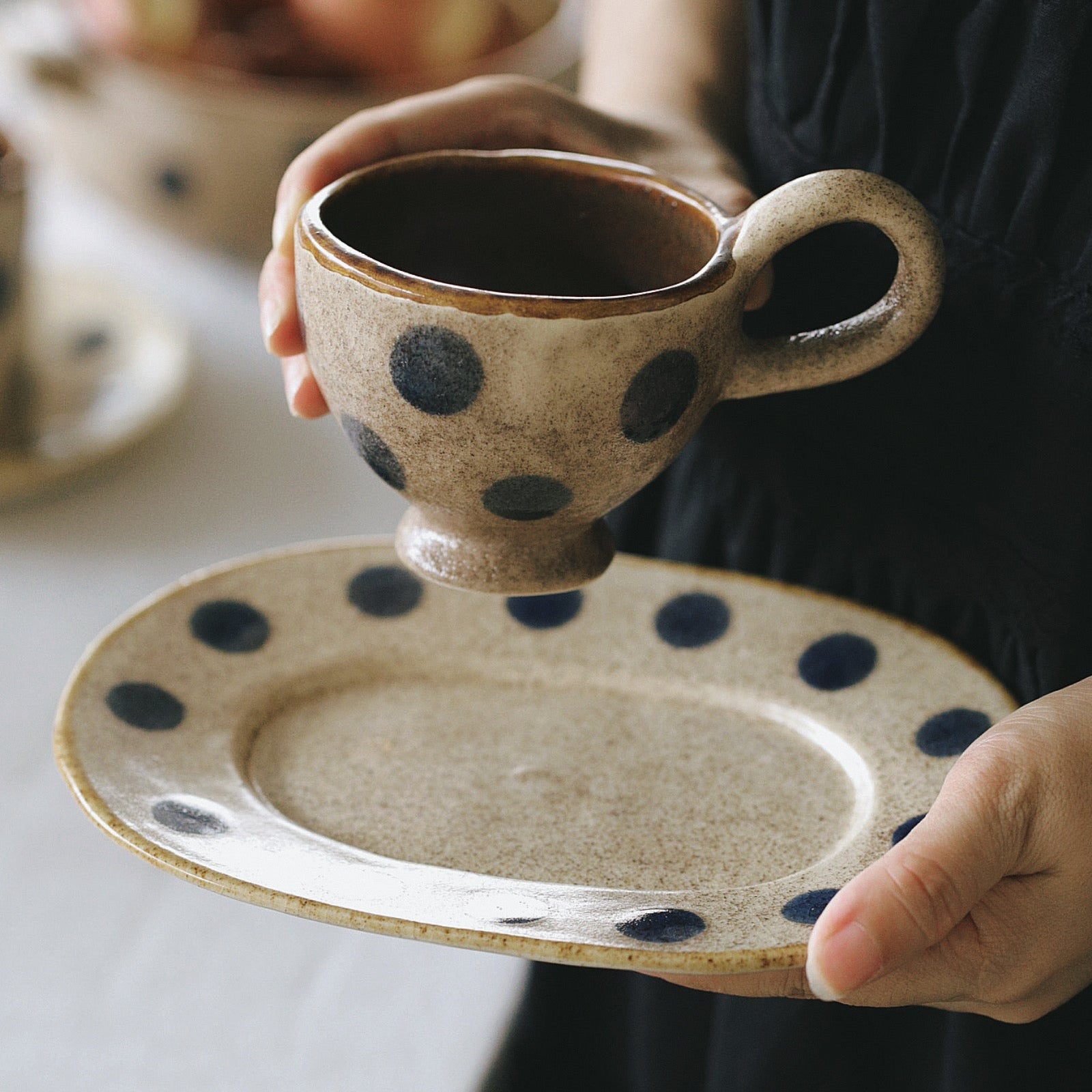 Person holding a ceramic cup and saucer with black polka dots on a neutral background
