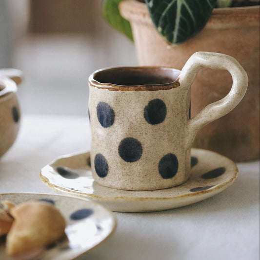 Ceramic cup with blue polka dots on a saucer, placed on a table with a plant in the background.